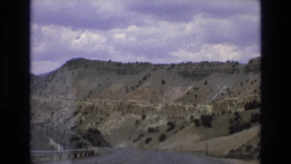 1973 8mm frame showing a downhill highway view through layered Nevada hillside terrain, with sparse shrubs, a roadside barrier, and heavy clouds above.