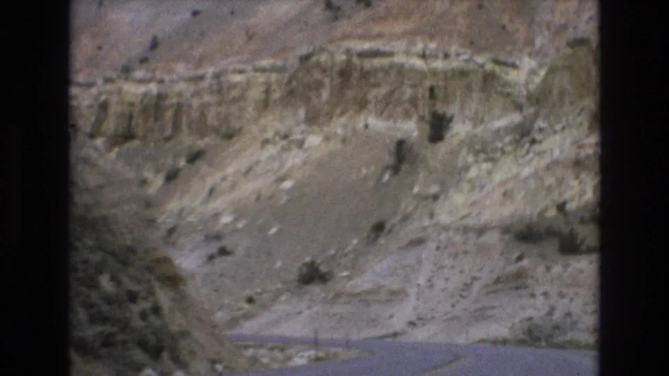 1973 8mm travel frame showing a first-person view of a winding road through arid canyon terrain near Lake Tahoe, Nevada, with eroded rock faces and scattered shrubs.