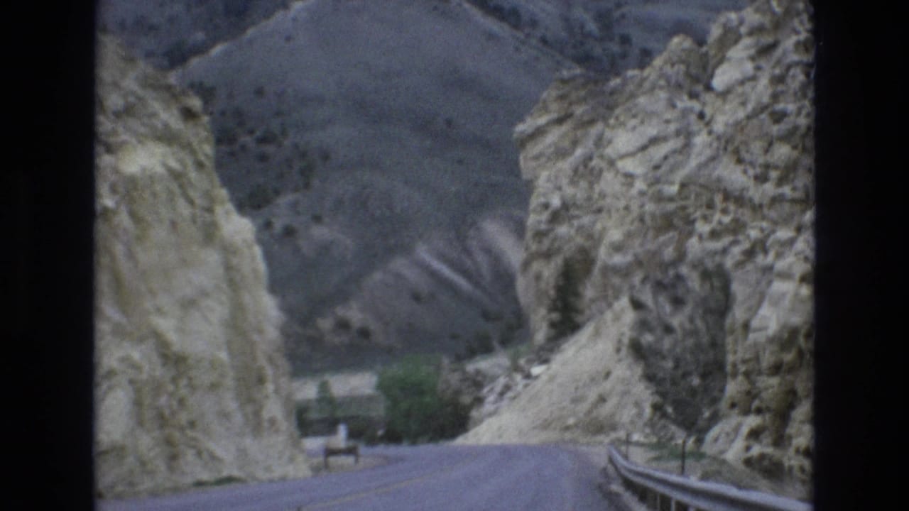 Curving road with guardrail beside rugged cliffs and distant hills.