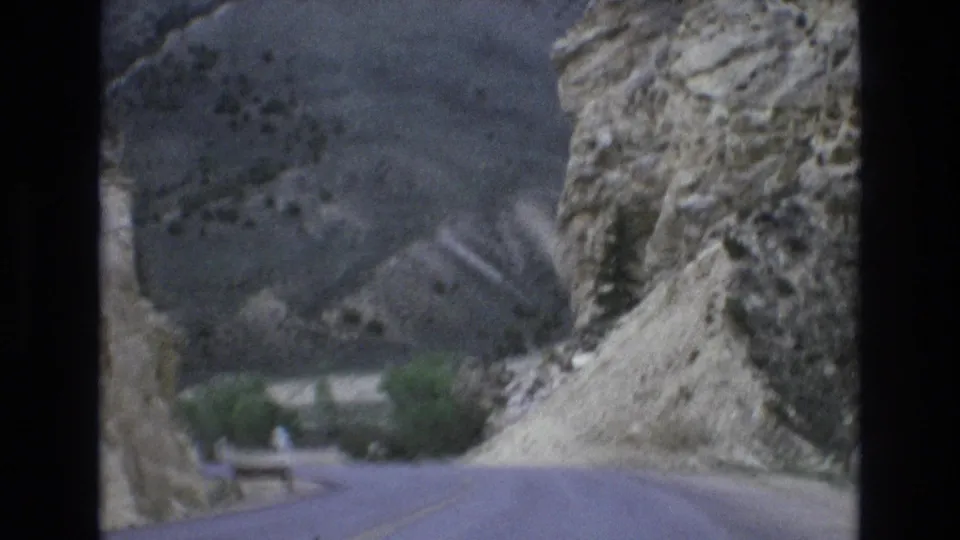 1973 Lake Tahoe, Nevada home-movie frame of a road curving past a rocky cliff, with a guardrail and green trees near the bend.