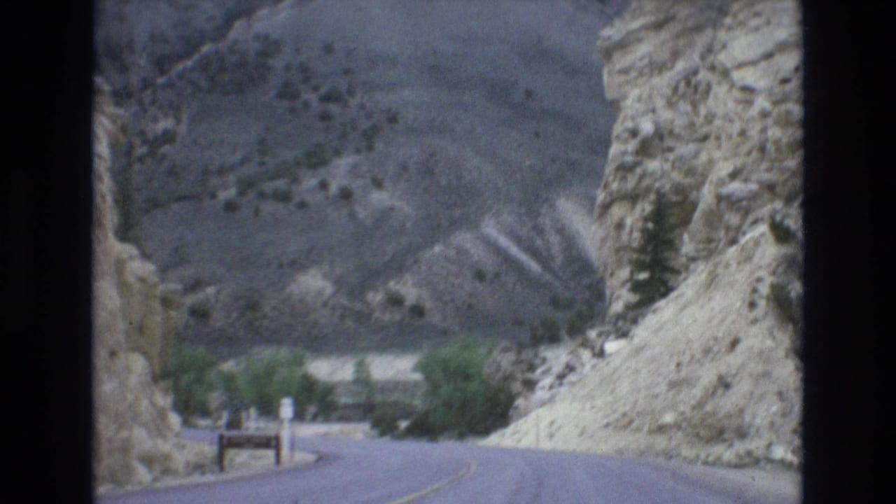 1973 8mm Lake Tahoe area road-view frame showing a paved road passing through rocky mountain terrain with sparse vegetation and a distant bend.