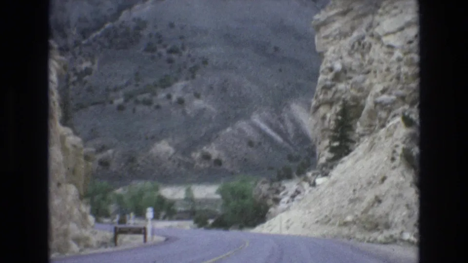 1973 8mm Lake Tahoe area road-view frame showing a paved road passing through rocky mountain terrain with sparse vegetation and a distant bend.