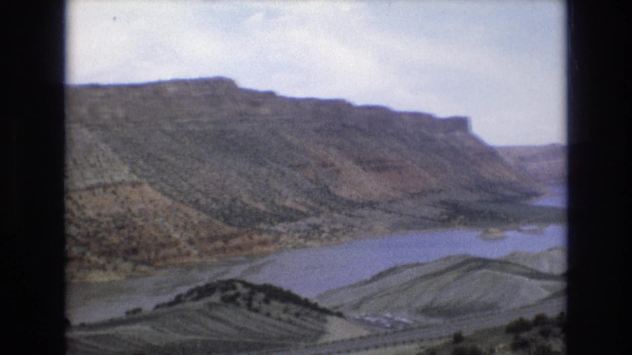 Wide canyon landscape with water below layered hills and sparse vegetation.