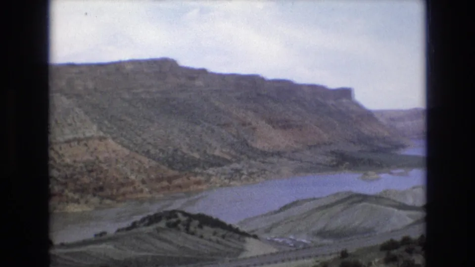 1973 8mm home-movie frame of a Lake Tahoe, Nevada-area canyon or lakeside overlook with layered rock slopes, sparse brush, and water in the valley below.
