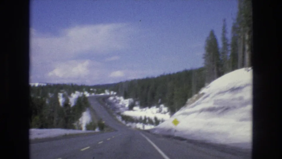 1973 Lake Tahoe, Nevada 8mm frame showing a paved mountain road receding between snowbanks and evergreen trees, with a yellow roadside sign and blue sky above.
