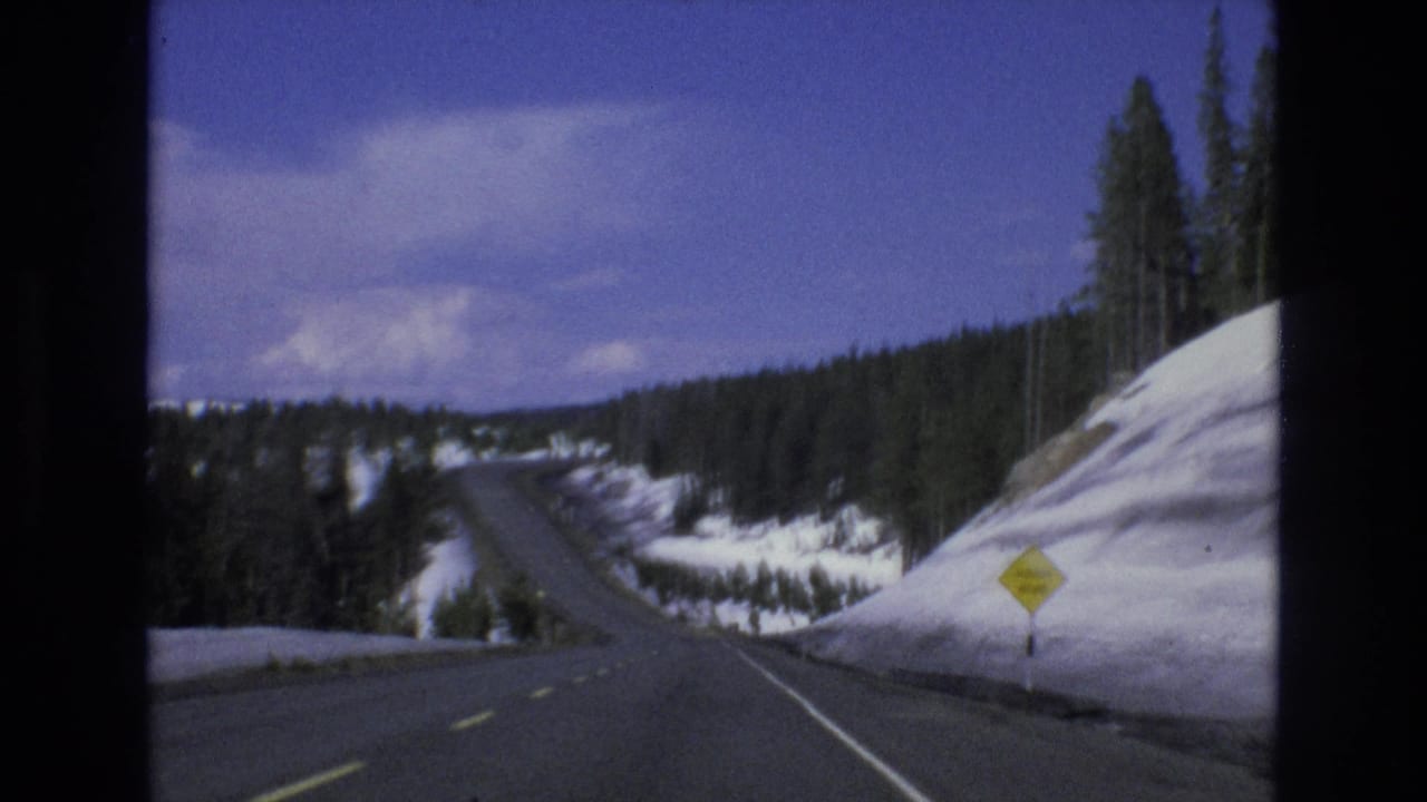 1973 Lake Tahoe, Nevada 8mm frame showing a curving two-lane mountain road with snowbanks, pine trees, and a yellow roadside warning sign.