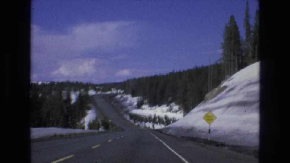 1973 Lake Tahoe, Nevada 8mm frame showing a curving two-lane mountain road with snowbanks, pine trees, and a yellow roadside warning sign.