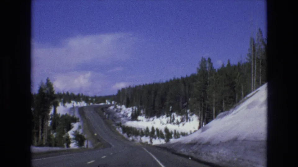 1973 Lake Tahoe, Nevada 8mm frame showing a vehicle-level view down a winding road with snow-covered shoulders and dense pine forest.