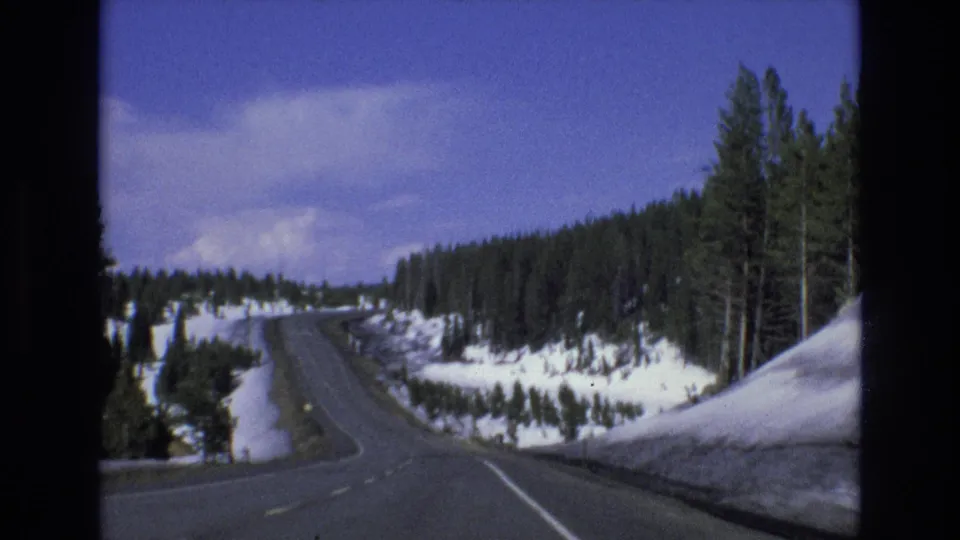 1973 Lake Tahoe, Nevada 8mm frame of a winding snowy mountain road passing between tall pines and banks of snow under a bright blue sky.