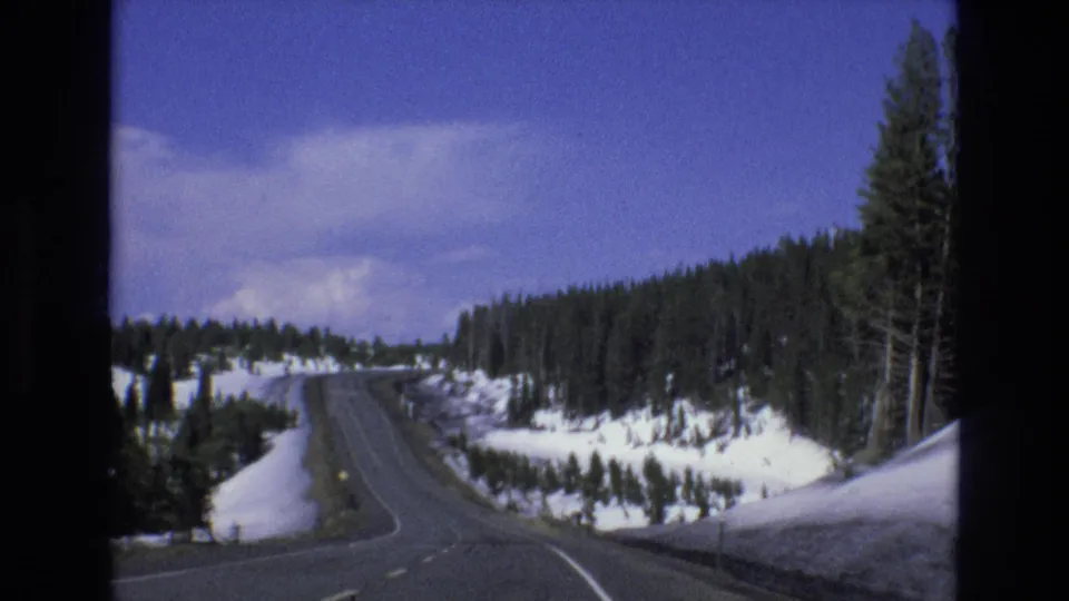 1973 8mm Lake Tahoe, Nevada car-perspective view of a two-lane mountain road curving through snow-covered evergreen terrain.
