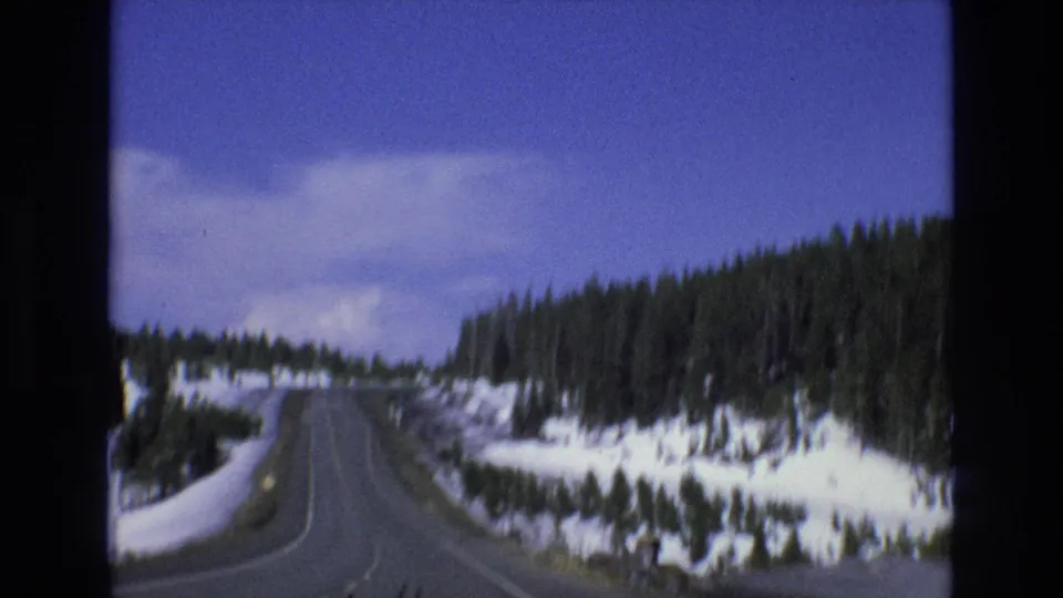 1973 home-movie frame from inside or near a moving car showing a snowy Lake Tahoe area road bordered by pine trees and bright sky.