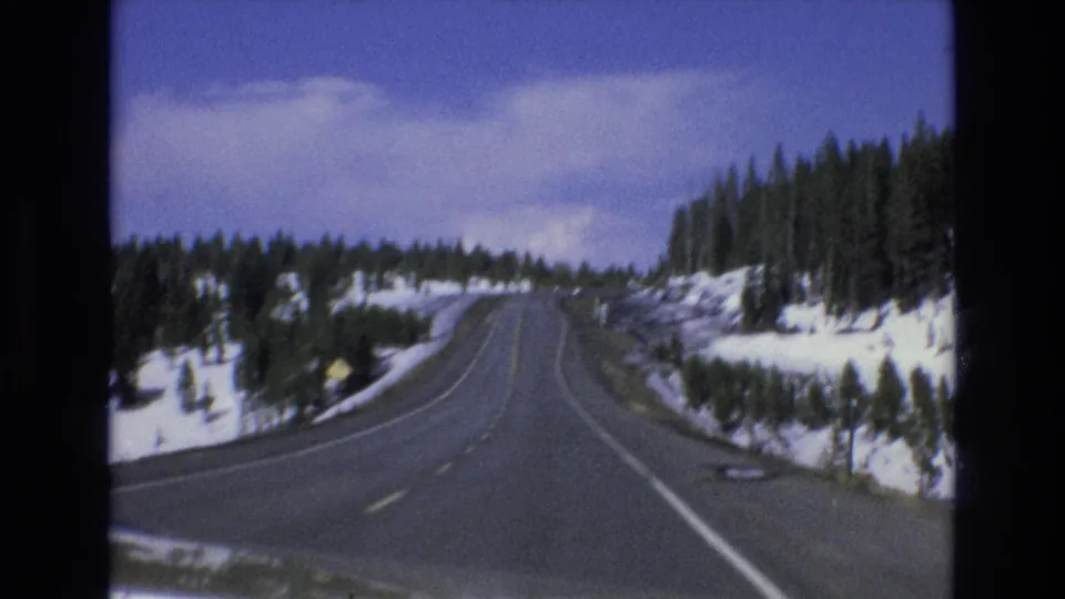 Vintage 8mm frame of a winding snowy mountain road near Lake Tahoe, Nevada, with evergreen slopes and an indistinct roadside sign visible in the distance.