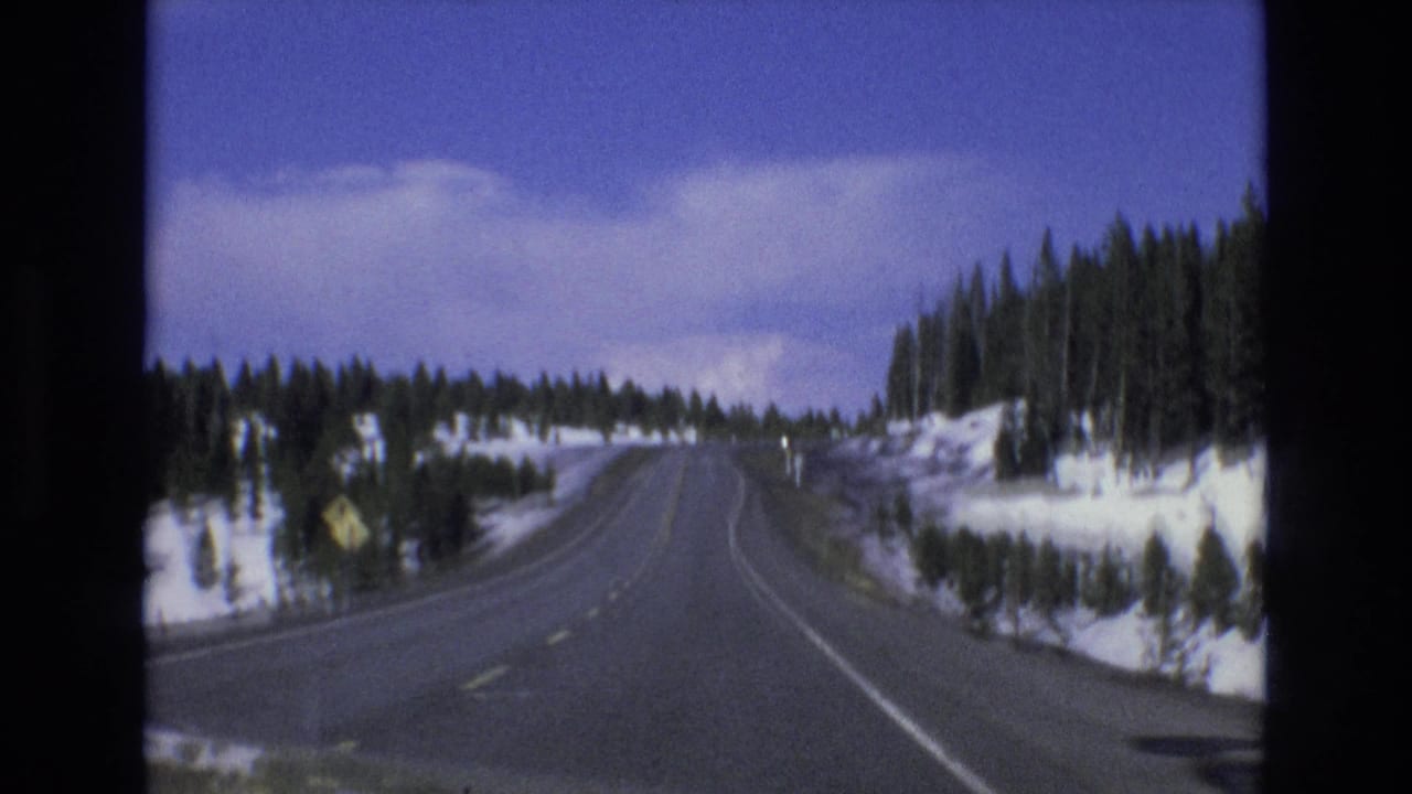 Snowy two-lane road curves through evergreen forest under a blue sky.