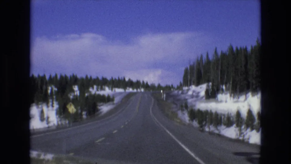 1973 Lake Tahoe, Nevada home-movie frame showing a winding snowy highway bordered by evergreen trees and snowbanks beneath a blue sky.
