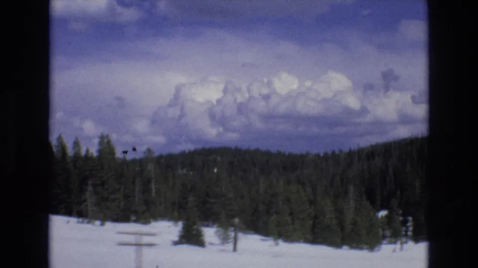 1973 Lake Tahoe, Nevada home-movie frame showing snow-covered ground, dark evergreen trees, utility poles, and a broad cloud formation.