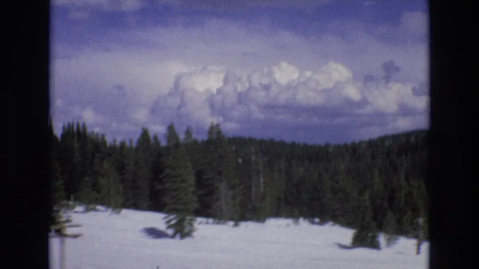 1973 Lake Tahoe, Nevada home-movie frame of snow-covered evergreen forest with utility poles and distant hills beneath a bright clouded sky.