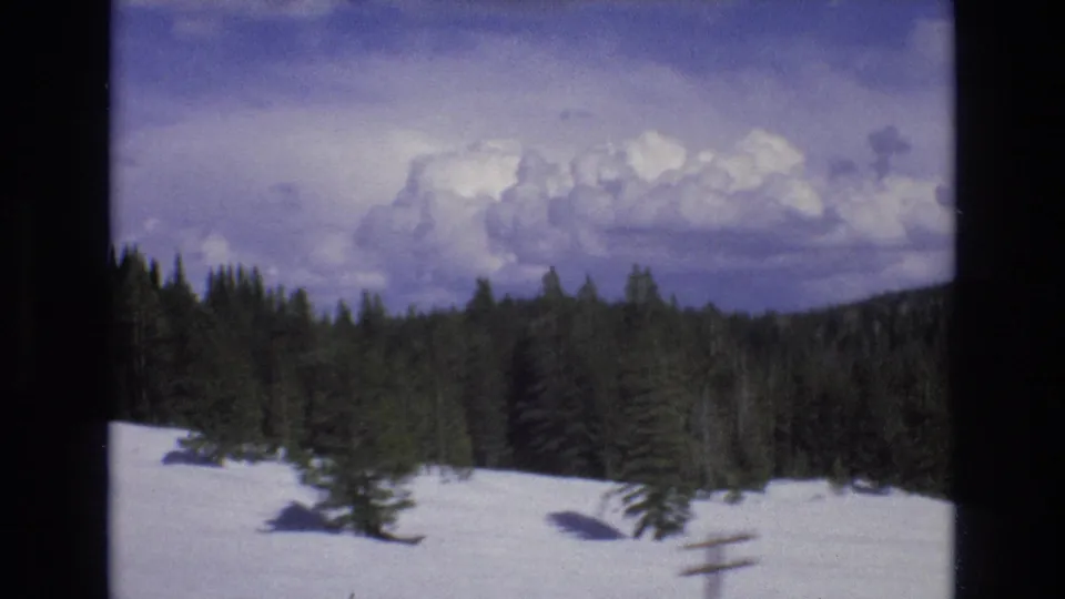 1970s 8mm frame of a snow-covered clearing bordered by evergreen forest beneath a cloudy mountain sky.