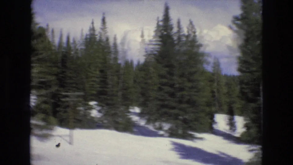 Handheld 8mm view across a snowy slope with dense evergreen trees and a pale clouded sky, consistent with a winter travel reel.