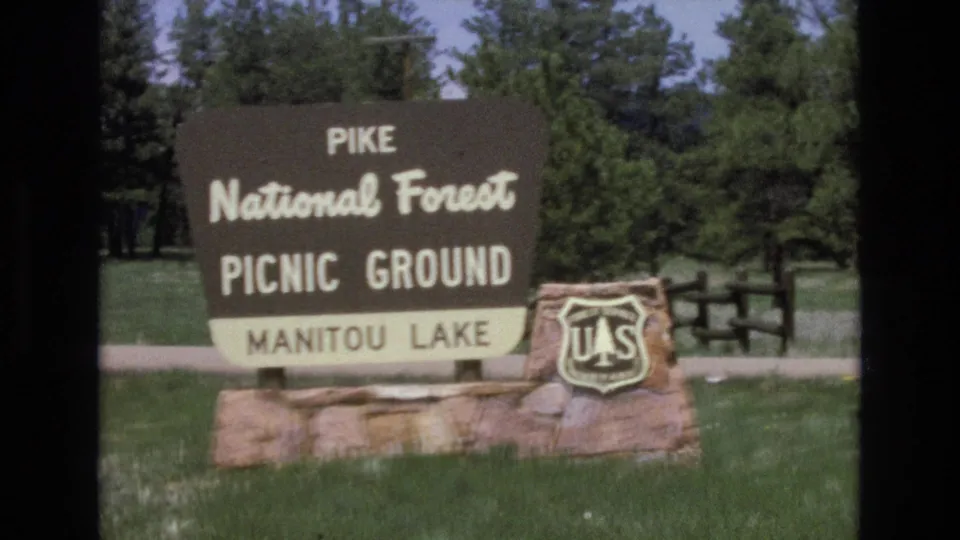 Visitor-shot 8mm frame showing a Pike National Forest Picnic Ground sign for Manitou Lake with a U.S. Forest Service shield beside grass, trees, and a fence.