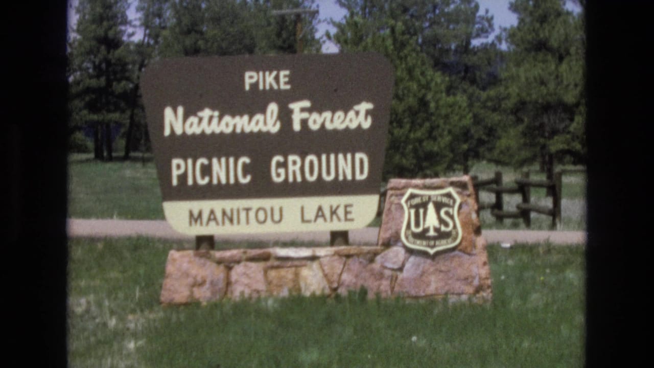 1970s 8mm frame showing a Pike National Forest Picnic Ground sign for Manitou Lake with a U.S. Forest Service shield on a stone base, backed by pine trees and grass.