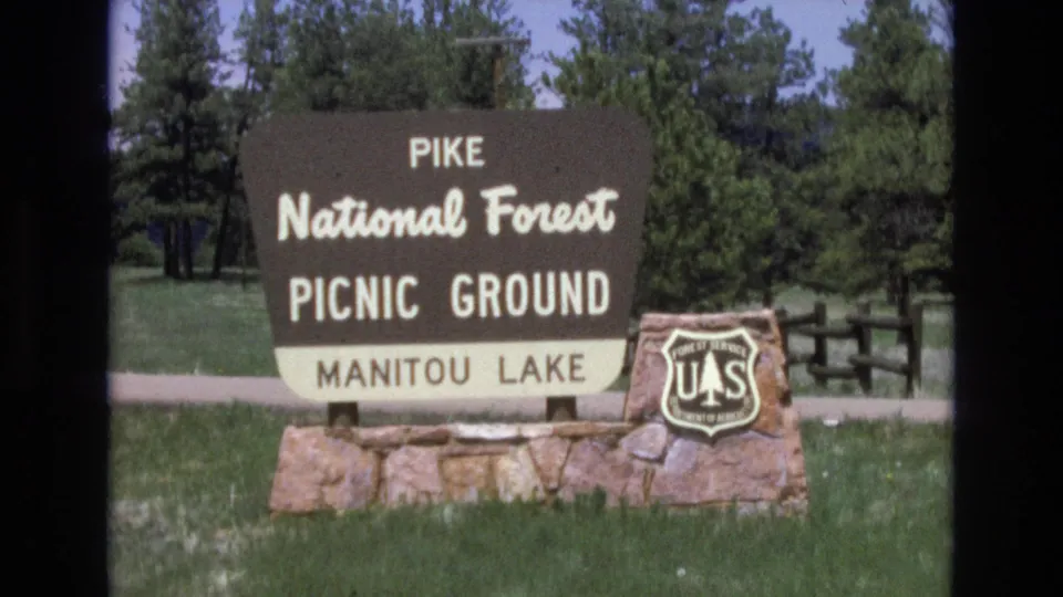 1970s 8mm still of the Pike National Forest Picnic Ground sign for Manitou Lake, framed slightly closer with the U.S. Forest Service shield, stone base, grass, and trees visible.