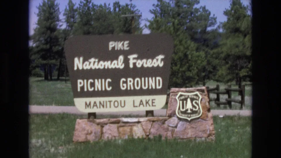 Final 8mm frame showing the Pike National Forest Picnic Ground sign for Manitou Lake from a steady visitor viewpoint, with the Forest Service shield, stone base, grass, wooden fence, and pine trees.