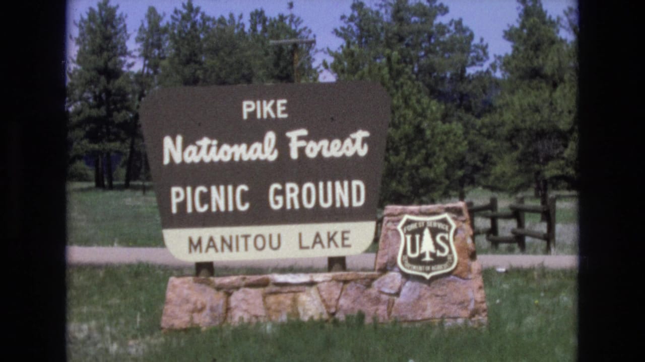 Pike National Forest picnic-ground sign for Manitou Lake beside trees and grass.