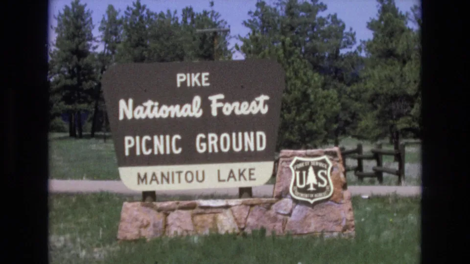 1970s 8mm frame showing a Pike National Forest picnic-ground sign for Manitou Lake with a U.S. Forest Service shield, grass, trees, and a road or path behind it.
