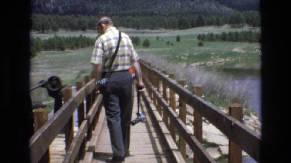 1970s 8mm frame of an adult man in a plaid shirt and dark trousers walking away on a wooden bridge at Manitou Lake, carrying a camera with green meadow, lake water, and trees beyond.