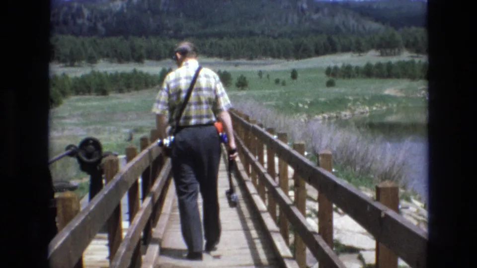 1970s 8mm frame showing the same adult man from behind on a wooden bridge, with bridge railings, a camera in hand, meadow grass, lake water, and trees in the background.
