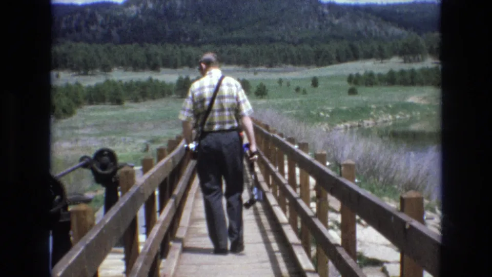 1970s 8mm frame of an adult man walking away down a wooden footbridge at Manitou Lake, with a camera, railings, open meadow, lake edge, and forested hills visible.
