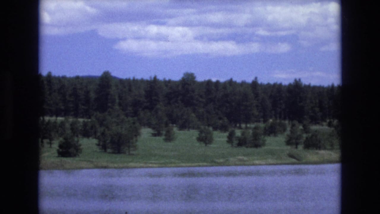1973 Lake Tahoe, Nevada home-movie frame showing calm water in the foreground, a green shoreline, dense trees, and a partly cloudy sky through dark film or window borders.