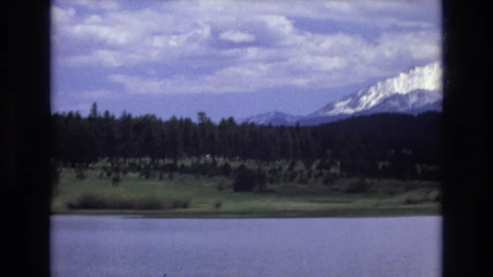 1973 Lake Tahoe, Nevada home-movie frame showing calm lake water, green shoreline, dark pine forest, cloudy sky, and a snow-capped mountain at right.