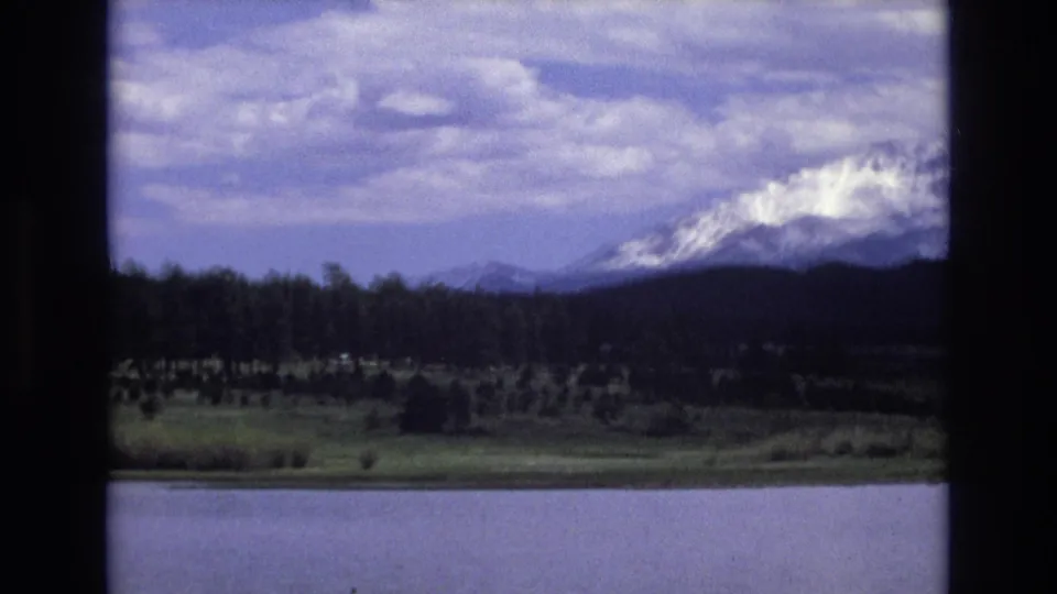 1973 8mm Lake Tahoe view across calm water toward a tree-lined shore and snow-capped mountains under a cloudy sky.