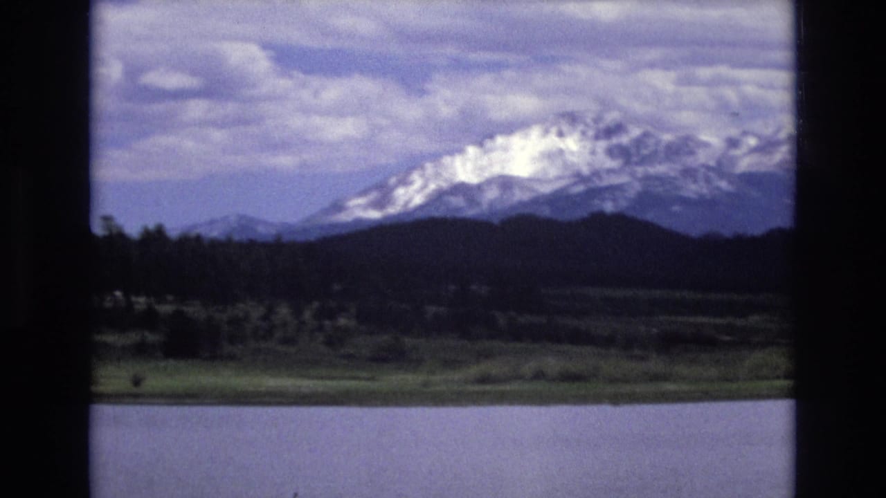 Clouds over a lake with a wooded shoreline and snow-covered mountain ridge in the distance.