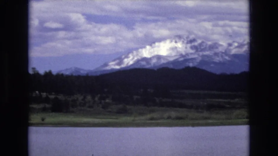 1973 Lake Tahoe scenery with calm foreground water, a dark evergreen band, and snow-covered mountains partly shaded by clouds.