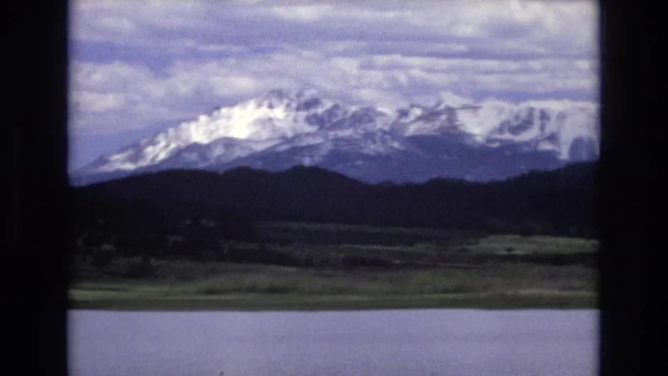1973 8mm frame showing Lake Tahoe water in front of a green shoreline, dark forest, and broad snow-capped mountain range beneath cloud cover.