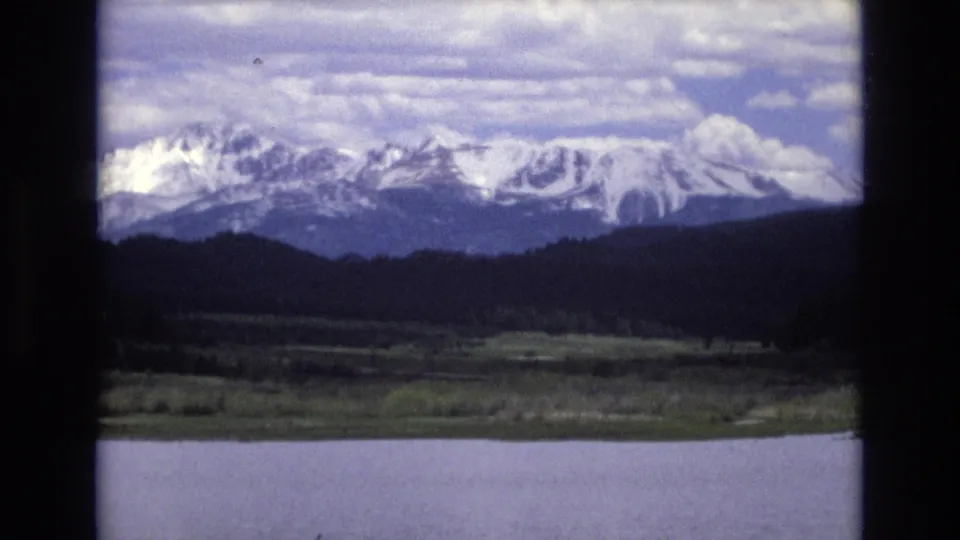 Wide 1973 Lake Tahoe home-movie view across the lake to a forested shore and snow-bright mountain peaks beneath a cloudy sky.