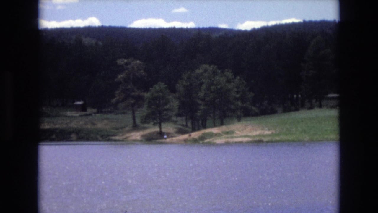 1973 Lake Tahoe, Nevada 8mm frame showing blue water in the foreground, a grassy shore, scattered trees, wooded hills, and a small distant structure.