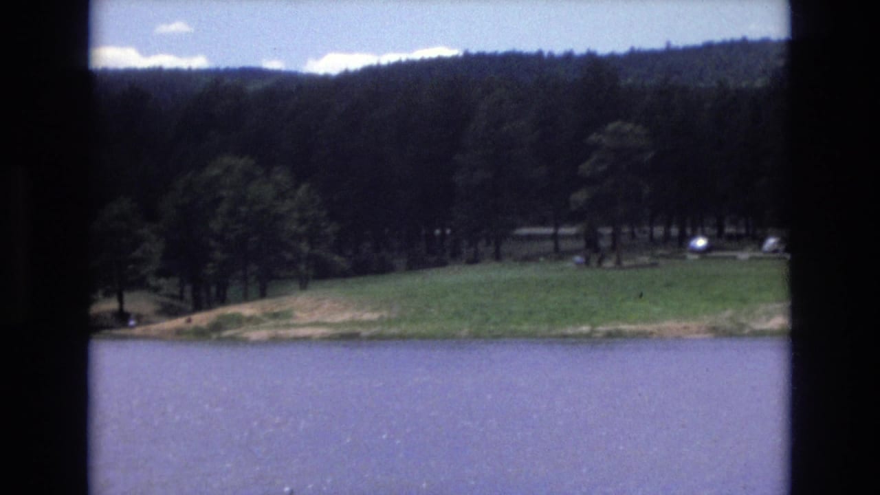 1973 Lake Tahoe, Nevada 8mm frame with lake water, a grassy shoreline, trees, wooded hills, and small distant vehicles near the trees.