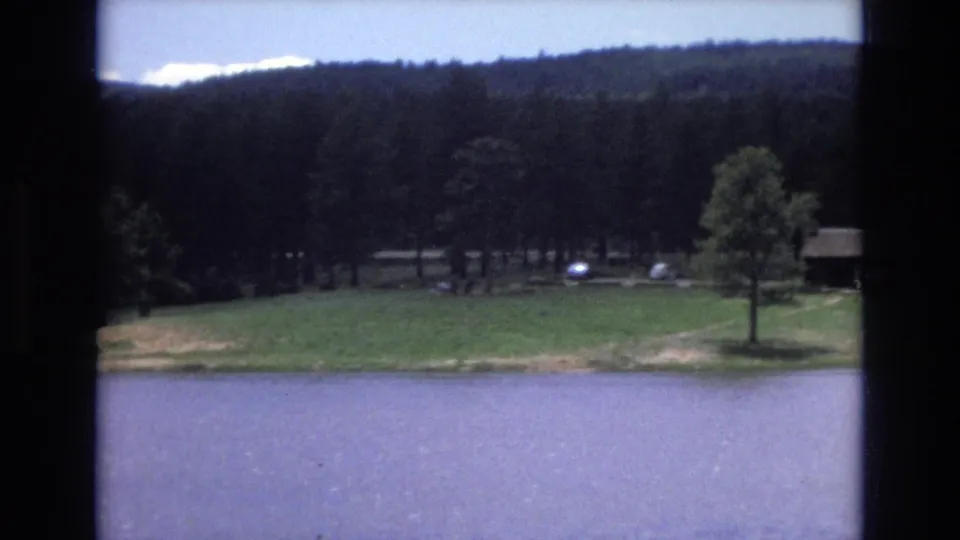 1973 Lake Tahoe, Nevada 8mm frame showing the lake edge, open grass, a prominent tree, distant vehicles, and a small cabin near the woods.