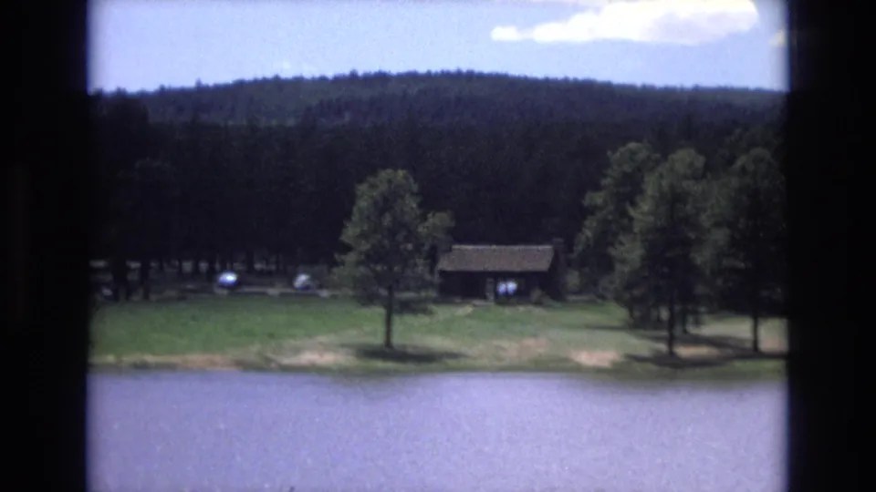 1973 Lake Tahoe, Nevada 8mm frame with blue water, grassy shoreline, a single tree, small wooden cabin, distant cars, and a forested hillside.