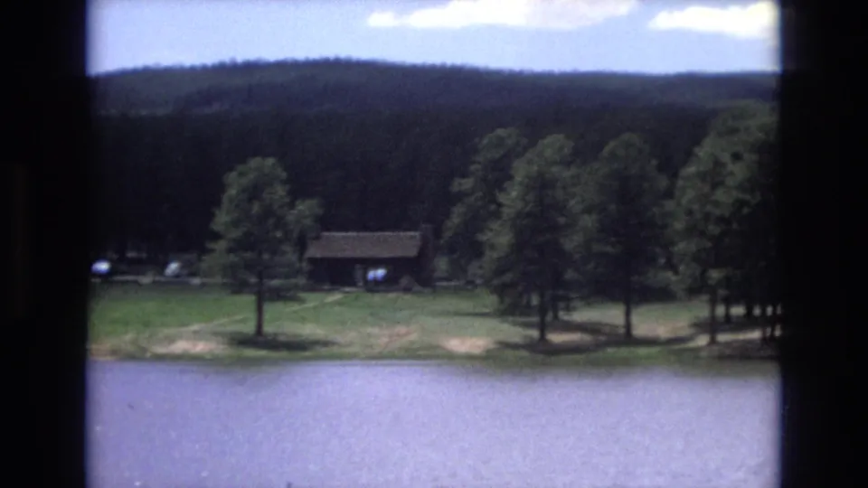 1973 Lake Tahoe, Nevada 8mm frame showing calm water, a grassy shore, scattered pine trees, and a small rustic building near the trees.