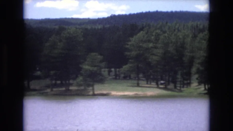 1973 Lake Tahoe, Nevada 8mm frame with calm water in the foreground, a sunlit grassy bank, tall pines, and a wooded hillside under clouds.