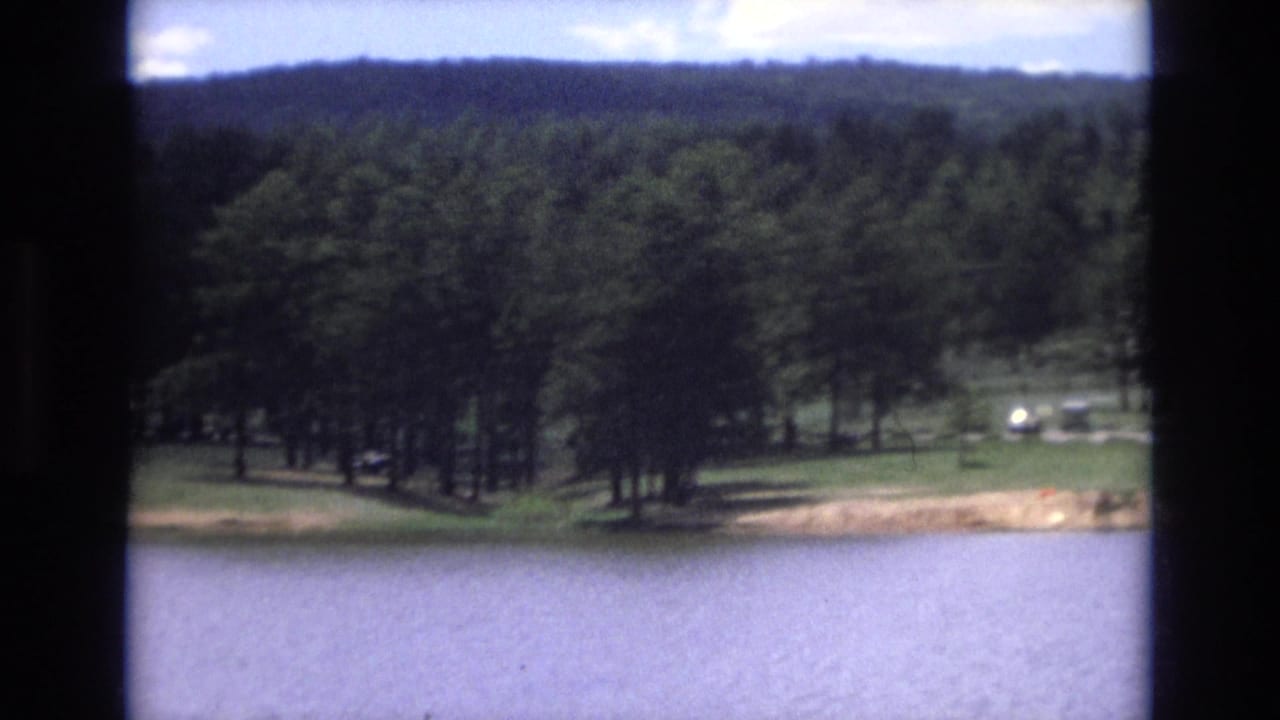 1973 Lake Tahoe, Nevada home-movie frame showing blue lake water, a narrow shore, tall pine trees, grassy lawn, and several small parked cars in the distance.