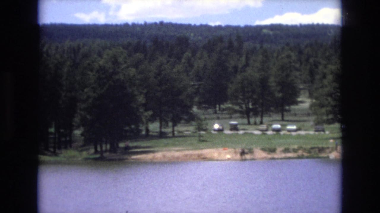 1973 Lake Tahoe, Nevada home-movie frame with calm lake water in front of a forested shoreline, a lawn, several parked cars, and tiny distant people near the water.