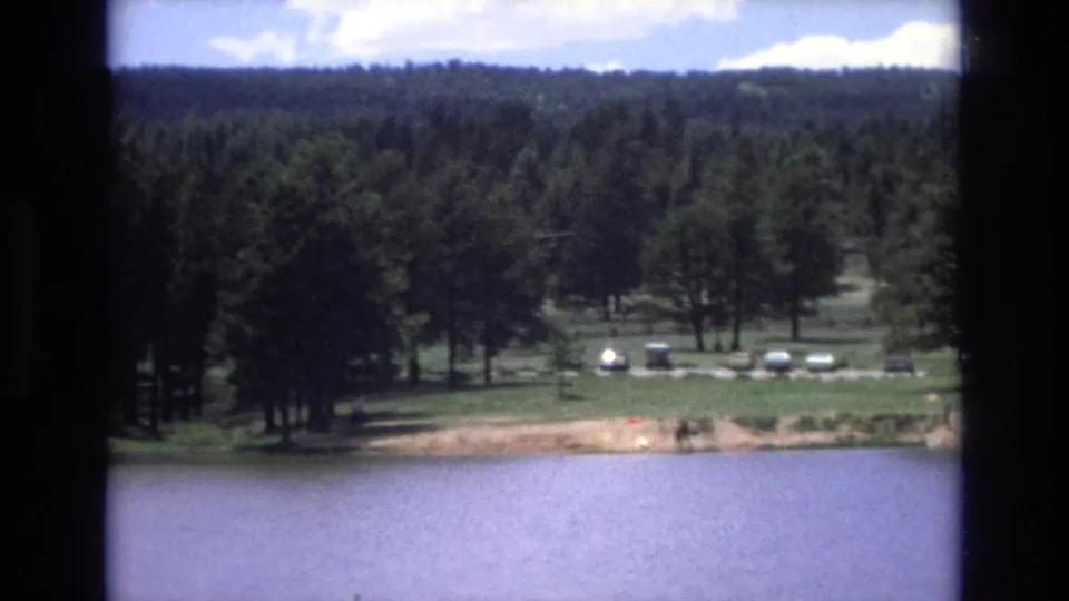 1973 Lake Tahoe, Nevada home-movie frame with calm lake water in front of a forested shoreline, a lawn, several parked cars, and tiny distant people near the water.