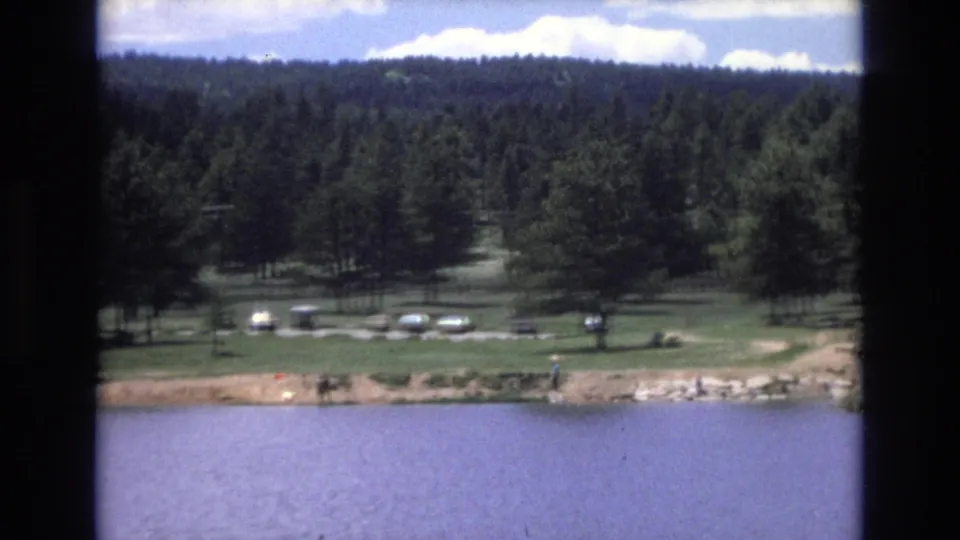 1973 Lake Tahoe, Nevada frame showing lake water below a shoreline recreation area with grass, tall trees, several parked cars, and small distant people along the edge.