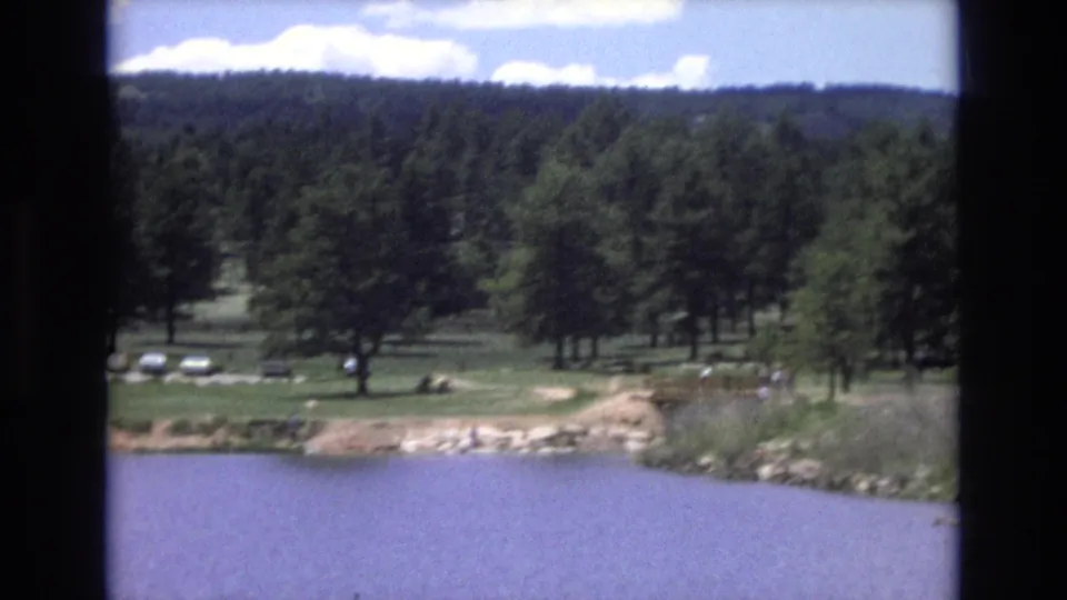 1973 Lake Tahoe, Nevada home-movie frame with blue water, rocky shoreline, a grassy area, tall pines, parked cars, and tiny people visible near the shore.
