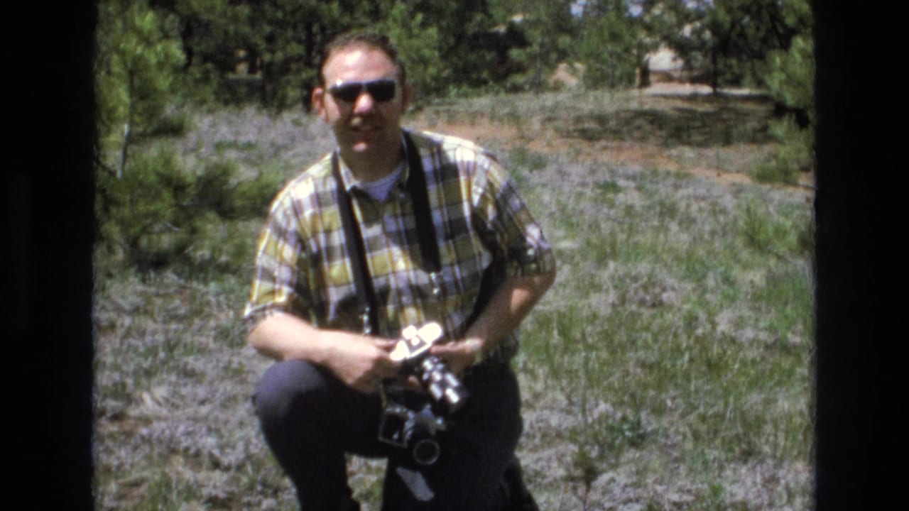 Adult man in a yellow and green plaid shirt and sunglasses crouches in a grassy Lake Tahoe park area while holding a camera with a strap.